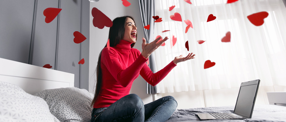 A woman sitting on her bed in front of her laptop throwing paper hearts