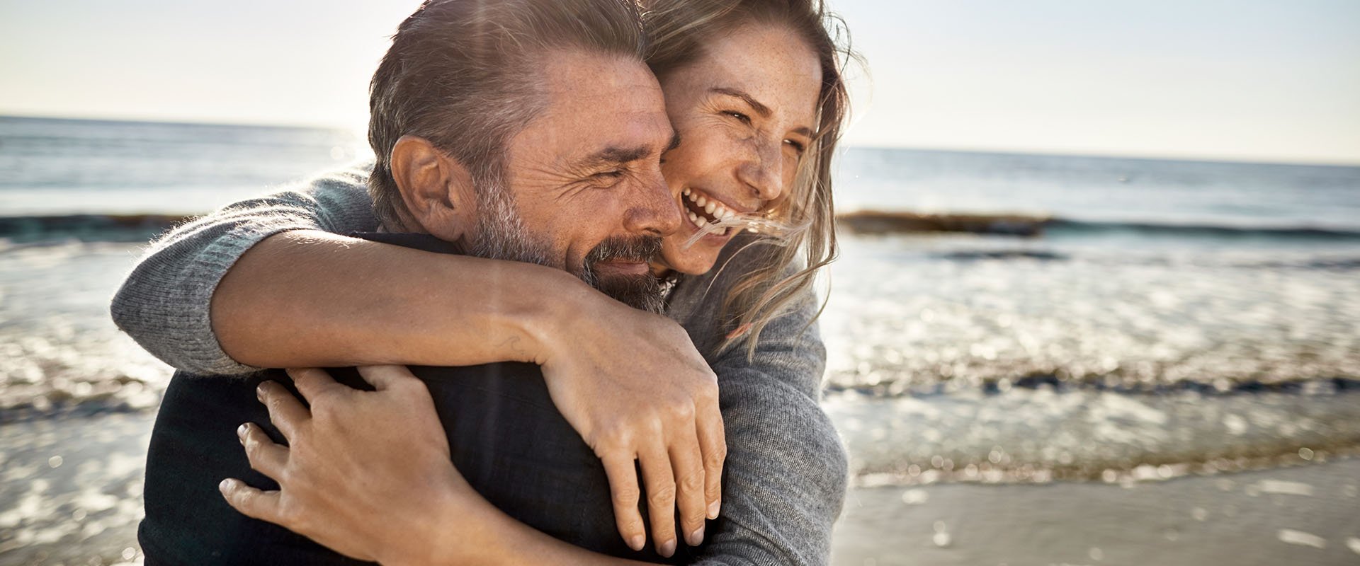 An age-gap-couple on the beach. They are hugging and laughing.