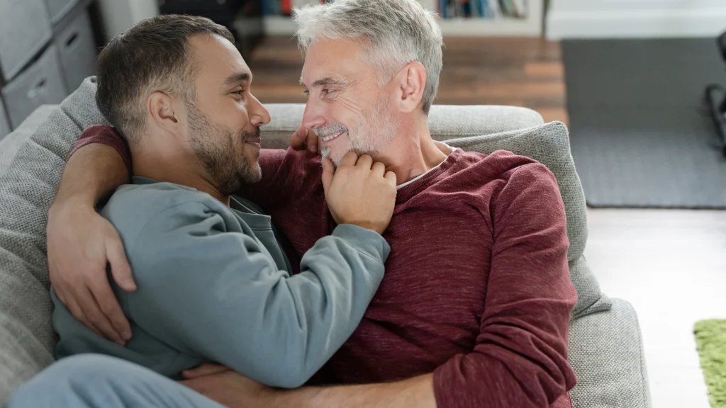 A gay couple over the age of 50 are cuddling together on their sofa.