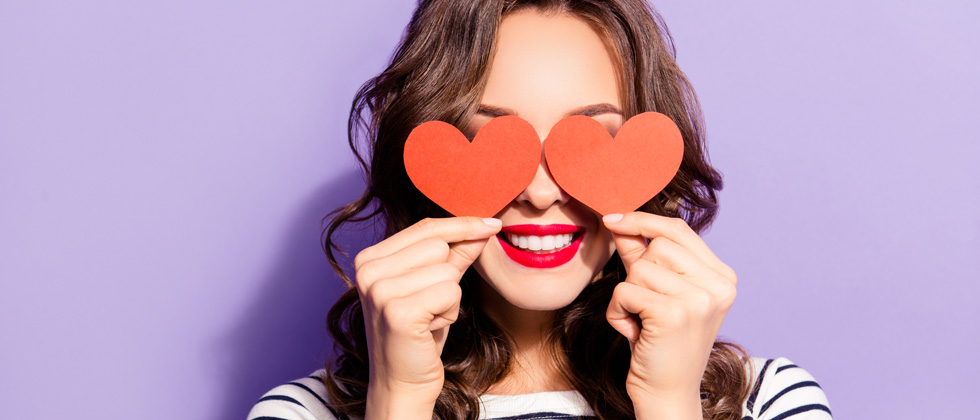 A smiling woman holding up two hearts over her eyes
