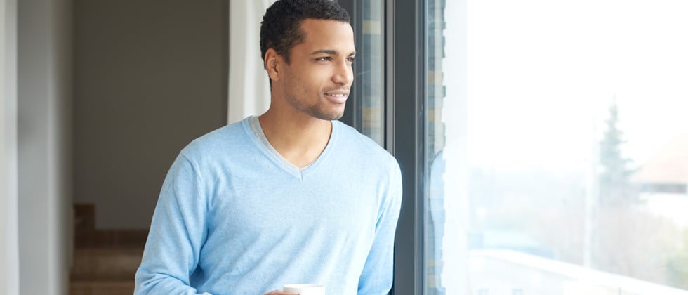 A young guy staring out a window holding coffee and smiling