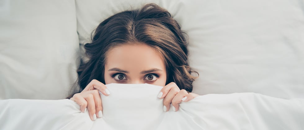 A woman in bed looking scared with the covers partly over her face