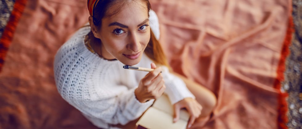 A young woman sitting on a picnic blanket writing in a journal