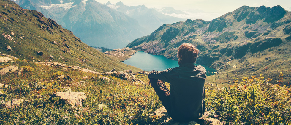 A guy sitting on a rock looking into a mountain valley with a lake at the base