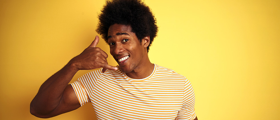 A young man stands against a yellow background, making a 'phone me' gesture with his right hand