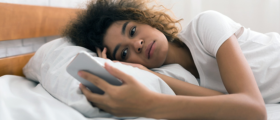 A young woman, looking troubled, lies in bed staring at her phone