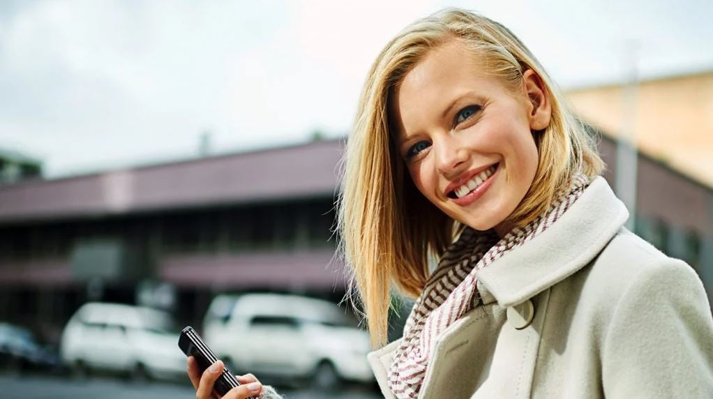 Woman with cell phone in hand smiling into the camera as a symbol for free dating at eharmony