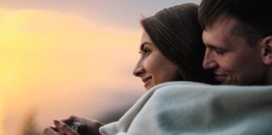 A couple cuddled under a blanket looking at the sunset outside