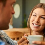 A woman and a man sitting next to each other. The woman has a cup in her hands and smiles at the guy.