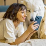 Woman sitting with her dog on the bed and looking to her phone.
