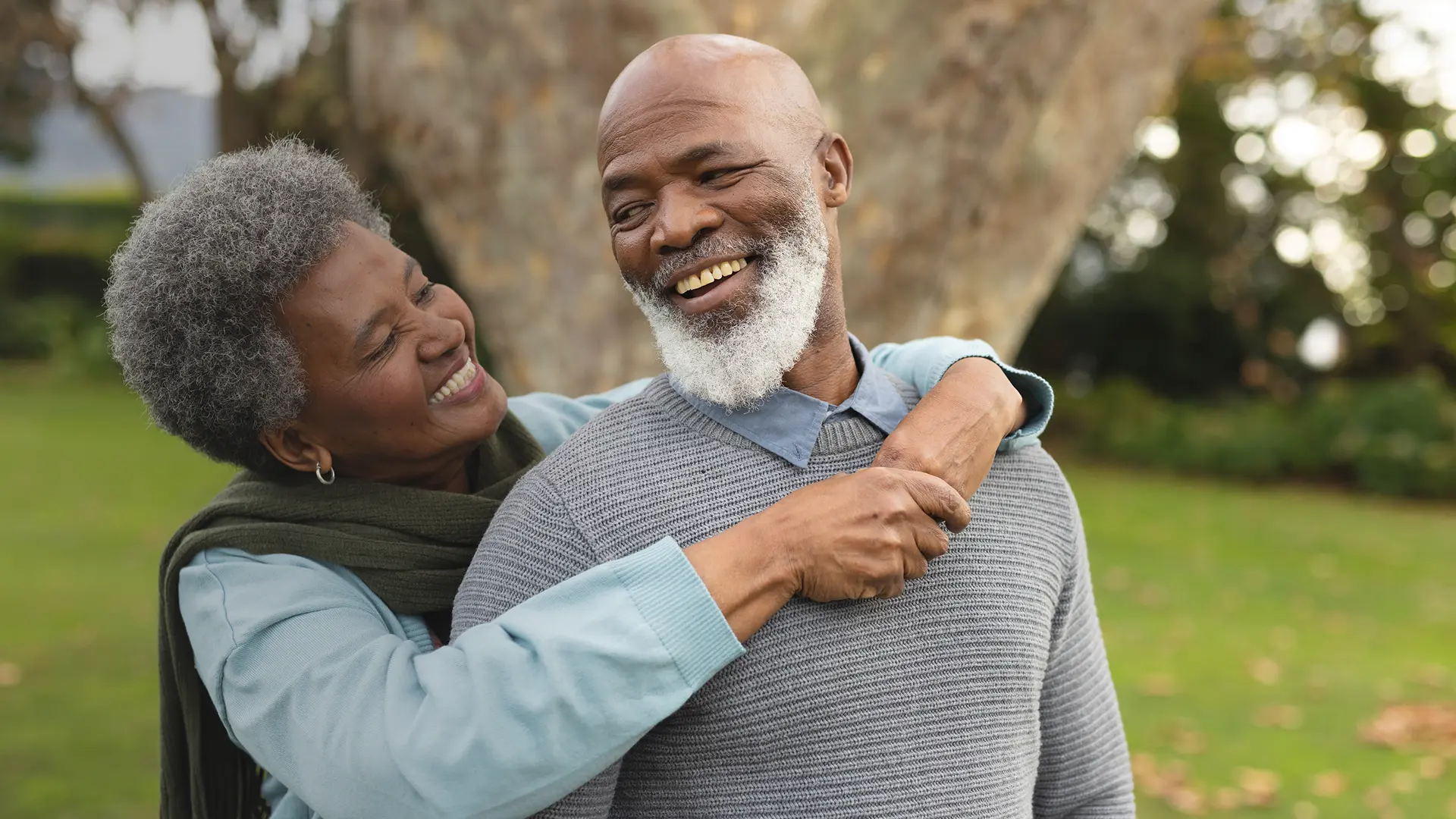 Black couple with the woman hugging her partner from behind in a park.